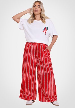 Kobieta o blond włosach, ubrana w białą koszulkę z motywem chili oraz szerokie spodnie w pionowe czerwono-białe paski, pozująca z ręką na głowie.