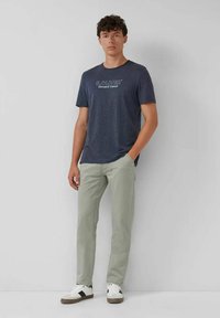 Junger Mann, der an einer Wand steht, trägt ein navy-blaues Grafik-T-Shirt, hellgraue Hose und weiße Sneaker mit schwarzen Akzenten.
