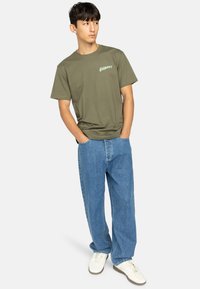 Olivgrünes Baumwoll-T-Shirt mit "ELEMENT" in hellgrün auf der Brust, kombiniert mit hellblauen Jeans und weißen Sneakern.