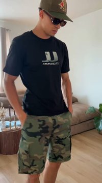 Man iklädd svart Jack & Jones t-shirt, kamouflageshorts, solglasögon och grön keps, står i vardagsrummet med soffa och växt.