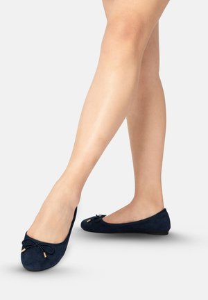 Jambes portant des ballerines en daim bleu foncé avec de petits nœuds et des bouts dorés, debout sur un fond clair uni.