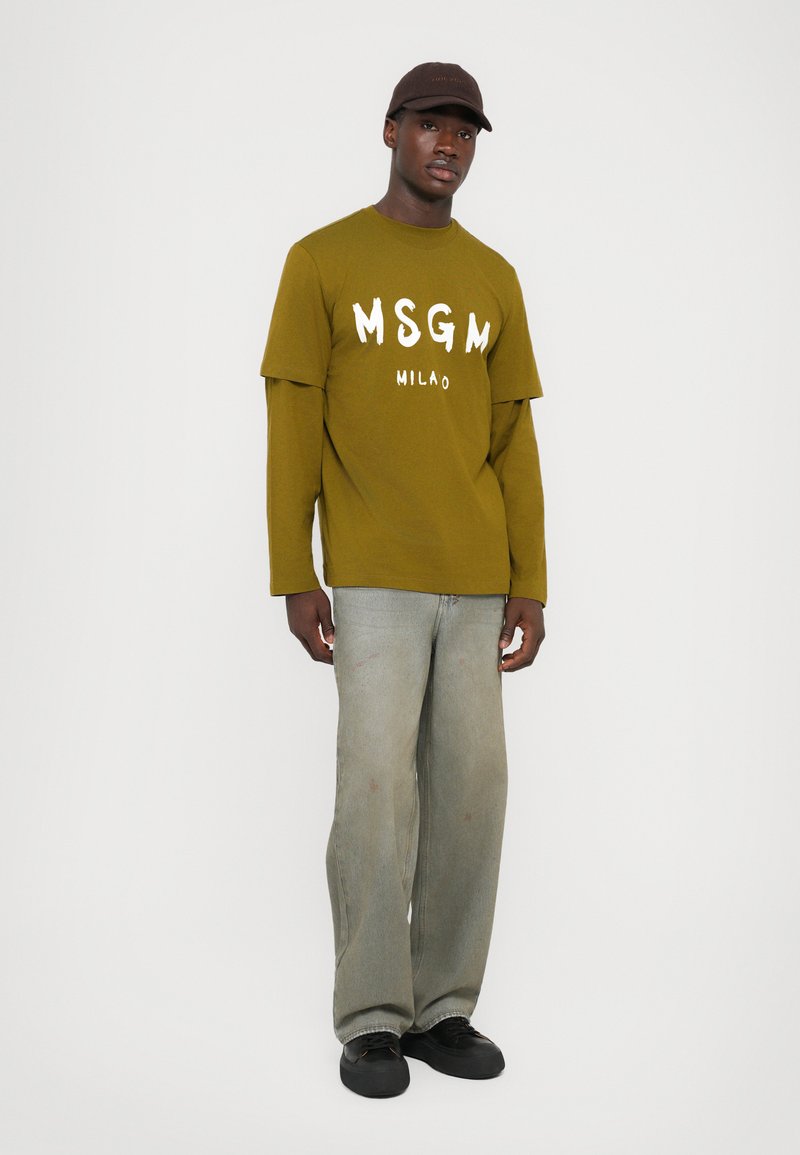 Man som bär en olivgrön långärmad lagertröja med texten "MSGM MILANO", ljus tvättade vida jeans, svarta sneakers och en mörkbrun keps.