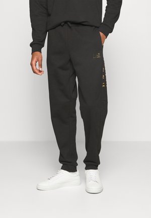 Mand iført sorte joggerbukser med guldlogo på låret og siden, kombineret med hvide sneakers, stående mod en ensfarvet baggrund.