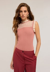 Top senza maniche ribbed rosa con collo alto e accenti testurizzati beige. Abbinato a pantaloni bordeaux con dettaglio dei bottoni.