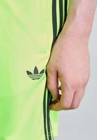 Neon green athletic shorts with black side stripes and an embossed black Adidas logo on the left leg. Smooth, lightweight fabric.