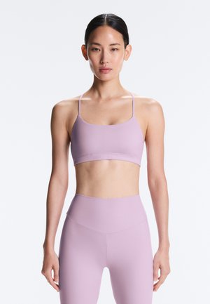 Vrouw met donker haar in een lage knot, gekleed in een bijpassende lichtpaarse sportbeha en hooggetailleerde legging, staand tegen een effen witte achtergrond.