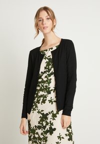 Cardigan nero realizzato in un tessuto liscio, con scollatura rotonda, maniche lunghe e chiusura a bottoni, indossato sopra un vestito a fantasia floreale.