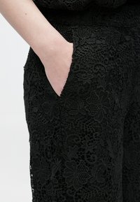 Χέρι που ακουμπά μέσα στην τσέπη μαύρου παντελονιού δαντέλας με κεντήματα λουλουδιών και περίπλοκες λεπτομέρειες από δίχτυ.