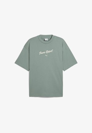 Miętowy, oversize'owy t-shirt z czarnym napisem, zestawiony z ciemnoniebieskimi spodniami typu wide-leg. Modelka trzyma torebkę w kolorze kremowym z falbanami.