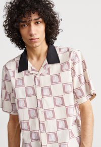 Short-sleeve shirt in cream fabric featuring a patterned design of purple and white stamps, with a black collar and button closure.