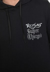 Sudadera negra con cordones que muestra texto en blanco de estilo gótico "Replay Super Charged" en el pecho.