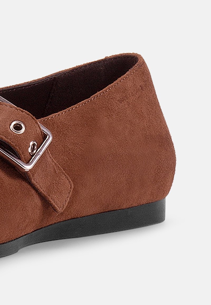 Braunes Wildleder-Mokassin mit abgerundeter Zehenpartie, ausgestattet mit einer silbernen Schnallenverzierung und einer niedrigen, schwarzen Gummisohle. Strukturierte Oberfläche mit glatten Kanten.