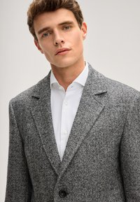 Blazer grigio a spina di pesce con rever a lancia, chiusura con un solo bottone e tessuto strutturato, abbinato a una camicia bianca con bottoni.