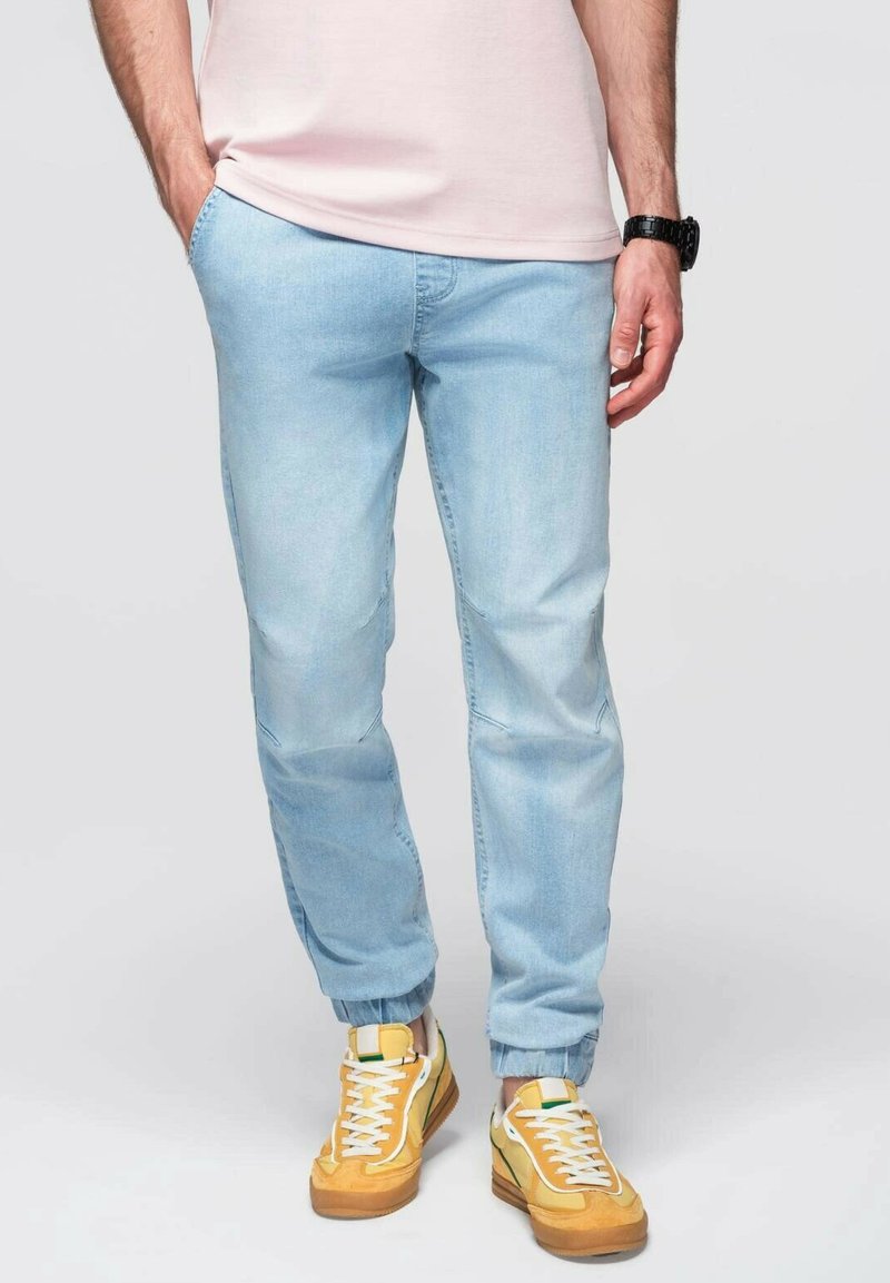 Světle modré džíny s tapered střihem, s bočními kapsami a manžetami, kombinované se žlutými teniskami a růžovým tričkem.