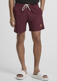 Burgunderfarbene Badeshorts mit elastischem Bund, Kordelzug und seitlichen Taschen. Mit einem Logo-Patch am linken Bein. Flauschige Textur und mittellange Beine bis zur Mitte des Oberschenkels.