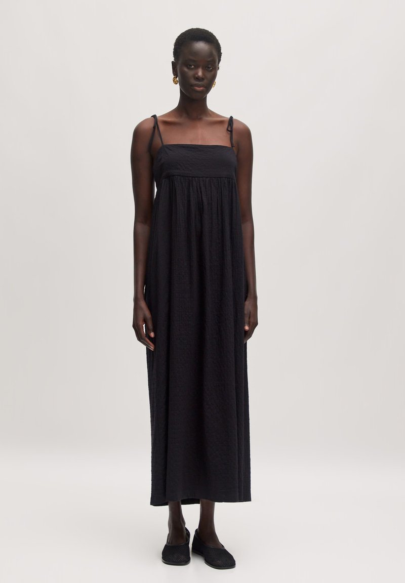 Schwarzes Maxikleid mit dünnen Trägern, strukturiertem Stoff und lockerer Passform, das bis zu den Knöcheln reicht. Schwarze Slipper.