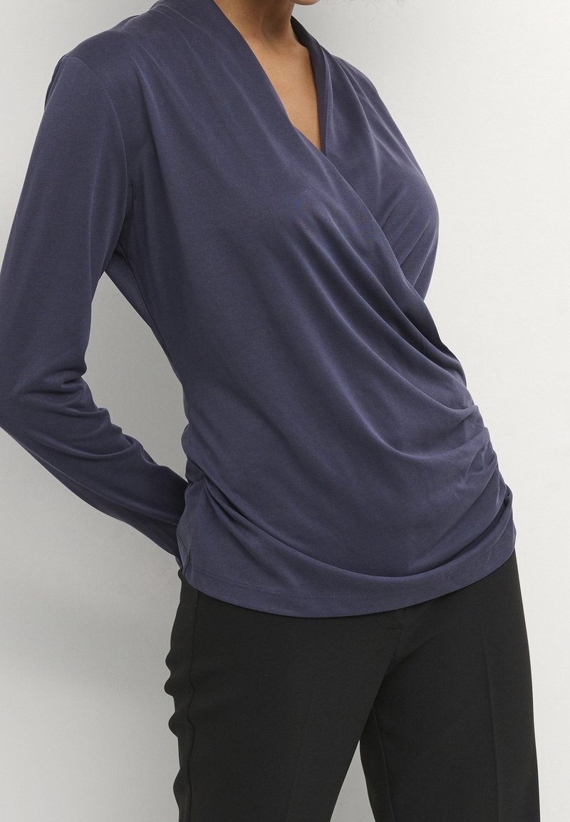 Kvinna iklädd en långärmad, marinblå inslagsblus med draperade sidor och svarta skräddarsydda byxor mot en enfärgad bakgrund.