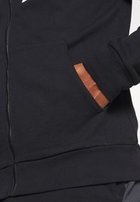 Hoodie noir zippé avec une texture de tissu lisse, comportant une poche avant et un accent taupe contrastant à l'intérieur de la poche.