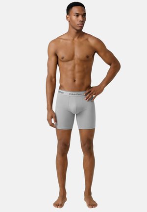Spiermatige man staat op blote voeten en draagt lichtgrijze Calvin Klein boxershorts met een gebrandmerkt elastiek, één hand op de heup, neutrale achtergrond.