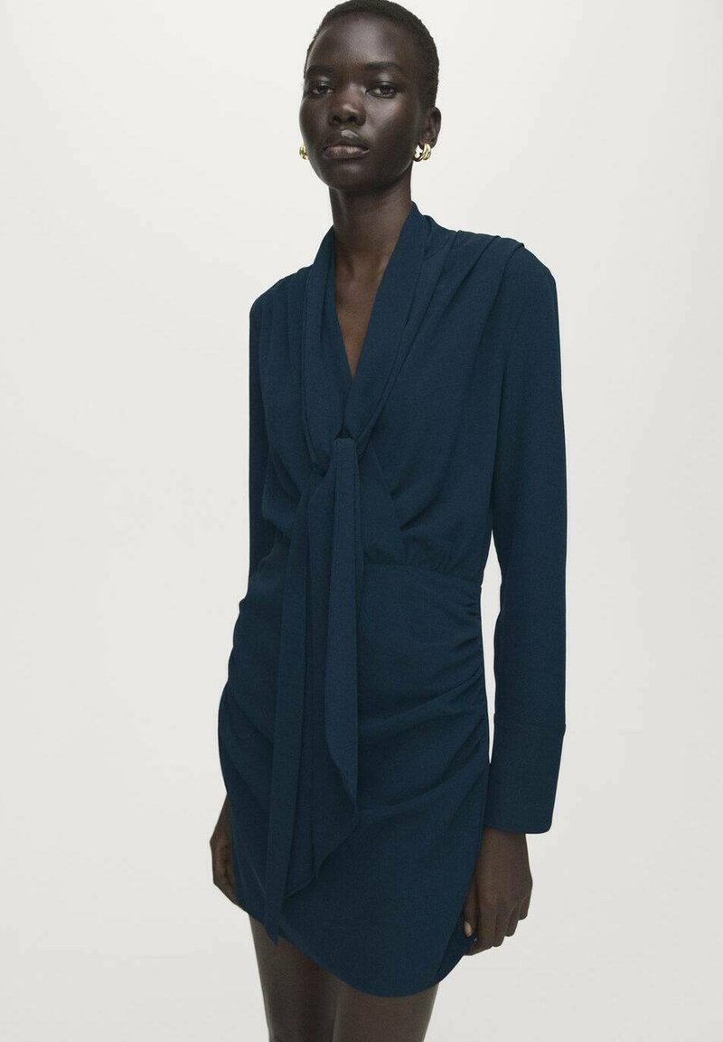 Marineblauwe jurk met lange mouwen, detail met strik aan de voorkant en geplooide stiksels in de taille, gemaakt van een gladde stof met een zachte textuur.