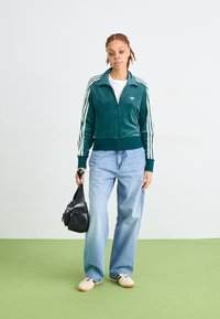 Pessoa jovem com uma jaqueta de treino azul-petróleo, jeans azul-claro e ténis, segurando uma bolsa preta, em pé sobre um chão verde, contra uma parede branca.