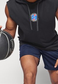 Schwarze ärmellose Kapuzenjacke mit einem Logo und grauen Kordeln, kombiniert mit navy-blauen Mesh-Shorts. Die Person hält einen schwarzen Basketball in einer Hand.