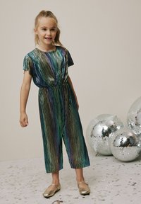 Tuta multicolore lucido con vestibilità ampia, maniche corte e gambe larghe. Il bambino indossa ballerine dorate; sfere disco sullo sfondo.
