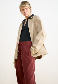 Beige Kapuzenjacke über einem schwarzen Shirt, kombiniert mit burgunderfarbenen, strukturierten Hose mit Kordelzug und Logo-Patch.