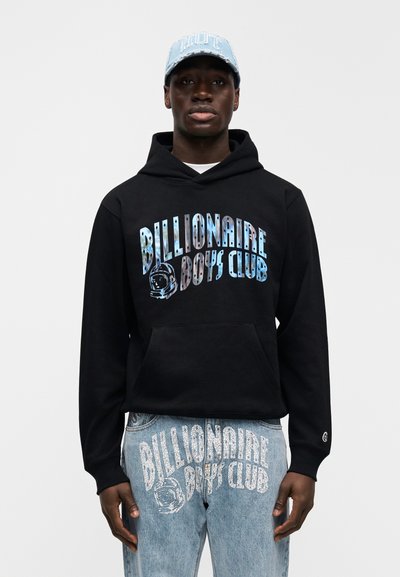 Μαύρο φούτερ με κουκούλα και μπλε γραφικό σχέδιο "Billionaire Boys Club" με σχεδιασμό αστροναύτη· συνδυασμένο με ανοιχτό μπλε τζιν παντελόνι με λευκή γραφή.