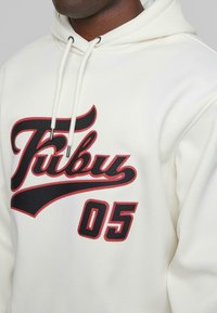 Hvit hettegenser med en stor svart og rød kursiv "Fubu"-logo og "05," med hvite snorer og teksturert stofffinish.