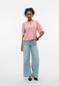 Donna con capelli ricci che indossa una blusa rosa morbida, jeans azzurri a gamba larga, scarpe con tacco scuro e un bracciale scuro, in posa contro uno sfondo bianco.