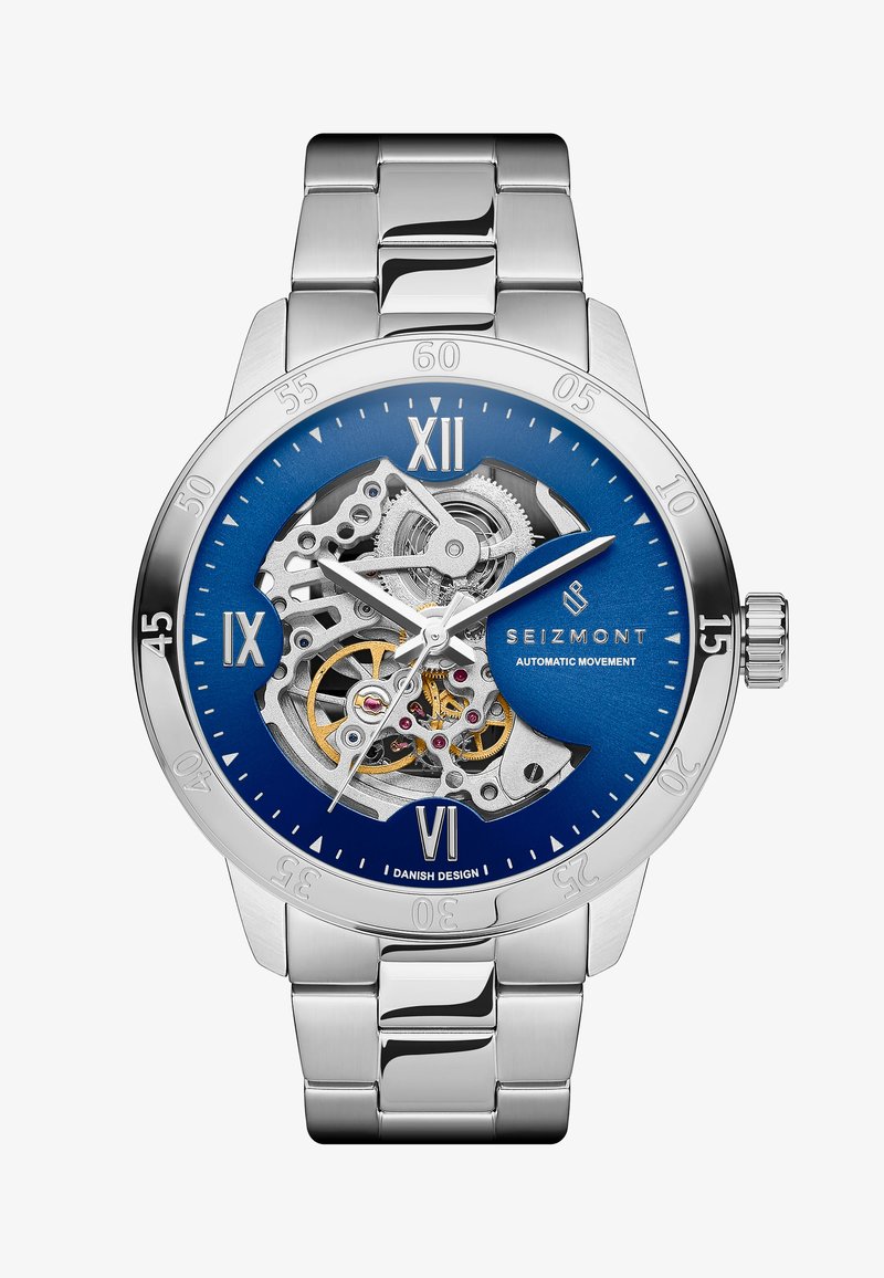 Rustfritt stål klokke med blå urskive, sølvfarget skjelettdesign, romerske tall og automatisk bevegelsesvisning. Børstet metallarmbånd.