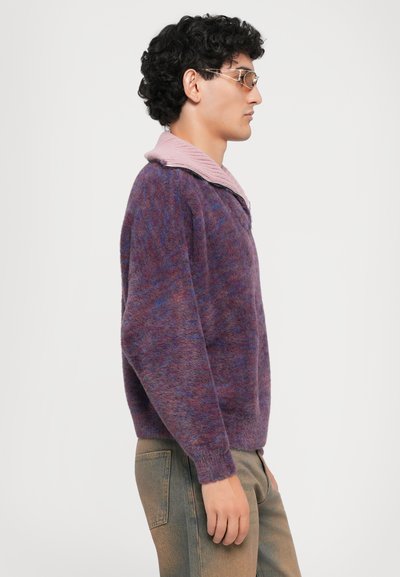 Purple and pink sweater with a textured finish, featuring a ribbed collar and a relaxed fit. Worn with light-colored, straight-leg jeans.