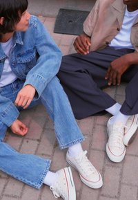 Deux personnes sont assises sur une surface en briques, portant des jeans en denim et des baskets décontractées. L'une porte une veste en denim ; l'autre a une chemise marron clair.
