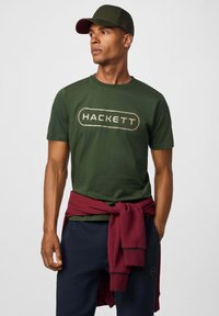 Camiseta de algodón verde oscuro con el logo "HACKETT" en dorado, combinada con un suéter burdeos atado a la cintura y pantalones azul marino.