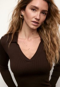 Brązowa prążkowana bluzka z długim rękawem i dekoltem w kształcie litery V. Luźne włókna tworzą teksturę. Złote geometryczne kolczyki dodają kontrastu. Naturalne, falowane włosy.
