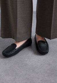 Mustat nahka loaferit, joissa on ommeltu kuvio, sileä pinta ja pyöreä kärki. Yhdistetty leveisiin, savisävyisiin housuihin.