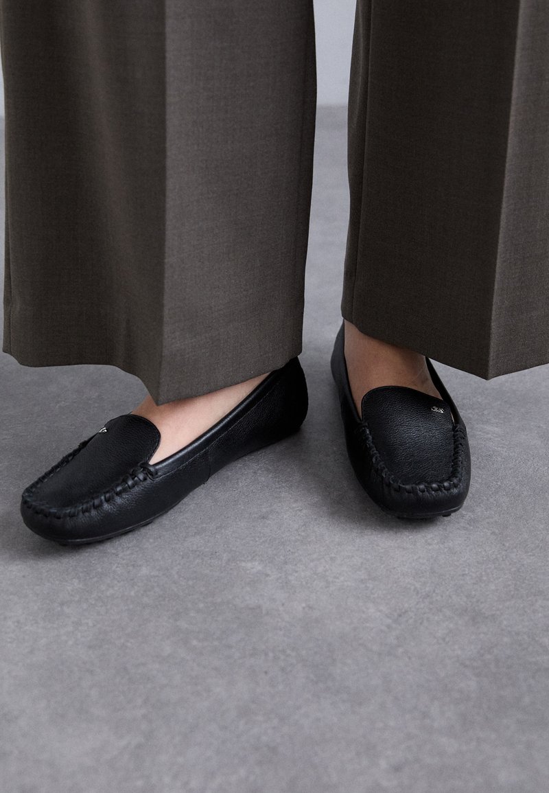 Mustat nahka loaferit, joissa on ommeltu kuvio, sileä pinta ja pyöreä kärki. Yhdistetty leveisiin, savisävyisiin housuihin.