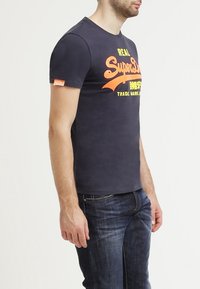 Námořnická modrá tričko s krátkým rukávem a kulatým výstřihem, na přední straně s živým oranžovým a žlutým grafickým textem. Měkký bavlněný materiál.