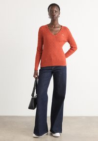 Orange cable-knit sweater with a V-neck, paired with dark blue wide-leg jeans and white sneakers, and a small black handbag.