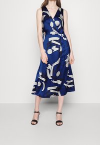 Blue wrap dress with abstract patterns in white and black, featuring a V-neckline and sleeveless design. Smooth fabric with a flowing silhouette.