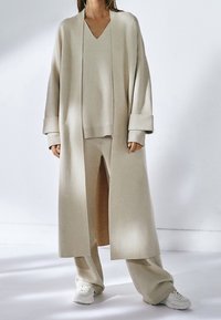 Manteau en tricot beige surdimensionné à manches longues, porté par-dessus un pull à col en V assorti et un pantalon à jambes larges, accompagné de baskets blanches.