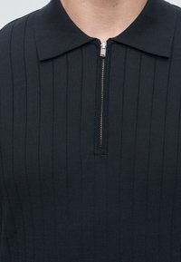 Gros plan d'une personne portant un pull en tricot côtelé noir avec une fermeture éclair argentée partiellement fermée au niveau du col.