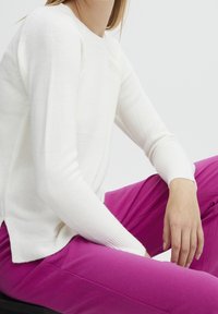 Personne assise, portant un pull blanc à manches longues et un pantalon rose vif, la main reposant sur le genou contre un fond blanc uni.