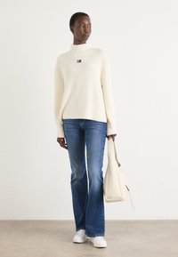 Cream ribbed knit sweater with a high neck, paired with blue flared jeans and white sneakers. Holds a beige shoulder bag.