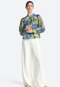 Bluza cu mânecă lungă cu imprimeu floral în verde, albastru și alb, cu decolteu adunat; asortată cu pantaloni largi albi și pantofi bej.