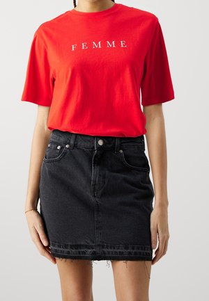 Femme portant un t-shirt rouge avec le mot "FEMME" et une jupe en jean noire taille haute avec des bords bruts.