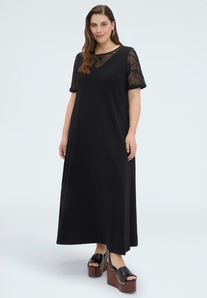 Frau trägt ein schwarzes Maxikleid mit transparenten floralen Spitzenärmeln und schwarzen Plateausandalen mit Holzabsätzen, steht vor einem schlichten Hintergrund.