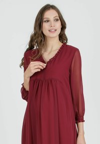 Burgundské šaty z lehkého materiálu, s výstřihem do V a volánkovým lemem, dlouhými průhlednými rukávy a staženým pasem pro elegantní siluetu.