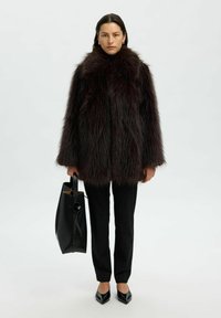Neppelsjas in donkerburgundy, met een hoge kraag en volumineuze mouwen. Draag het over zwarte broeken en houd een elegante zwarte handtas vast.